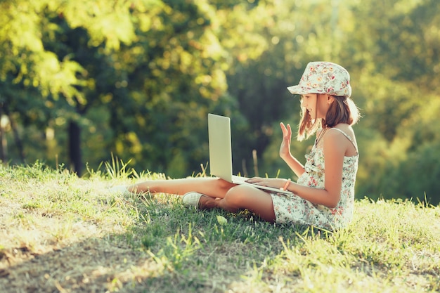 La bambina sta parlando su un computer portatile mentre era seduto sull'erba al sole. Vestito con un sarafan e un cappello