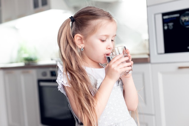 La bambina sveglia beve acqua fresca pulita da un bicchiere trasparente a casa in cucina proprietà utili dell'acqua per la crescita e lo sviluppo del bambino buone abitudini messa a fuoco selettiva