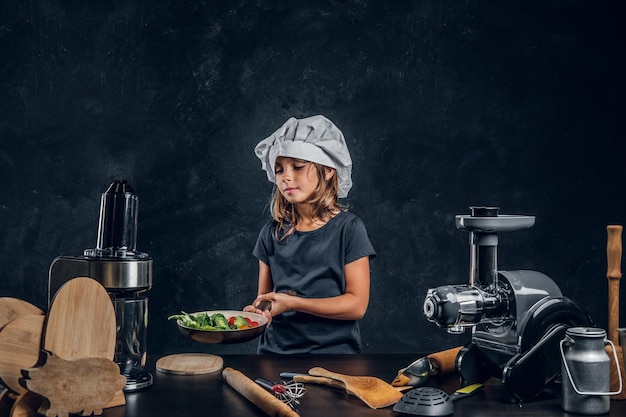 La bambina sveglia con il cappello da chef sta preparando le verdure per cucinare sullo sfondo scuro.