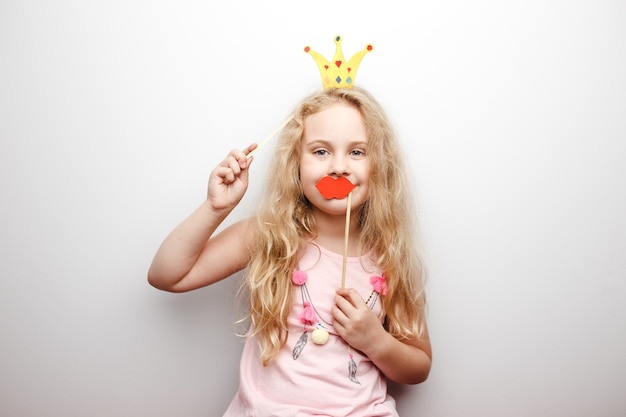 La bambina sveglia con la corona di carta e le labbra rosse sta sedendosi sulla sedia rossa a casa.