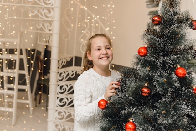 La bambina sveglia decora l'albero di Natale con i giocattoli di Capodanno e le palle rosse. Una ragazza in un maglione bianco e vestito si trova appeso palle su un abete artificiale