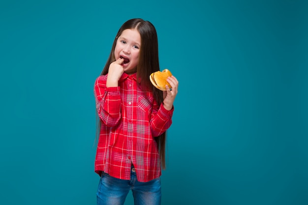 La bambina sveglia in camicia a quadretti con i capelli della brunetta tiene l'hamburger