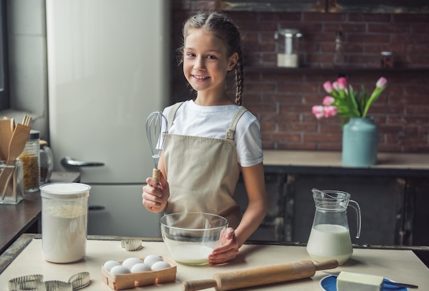 La bambina sveglia in grembiule tiene in mano una frusta guardando la fotocamera e sorride mentre prepara l'impasto in cucina a casa