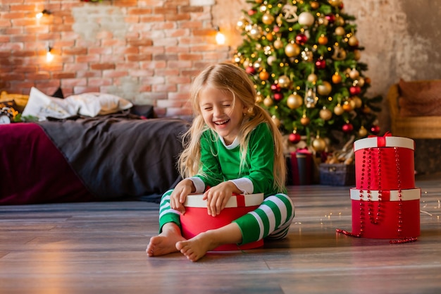 La bambina sveglia in pigiama vicino all'albero di Natale tira fuori il gattino della confezione regalo