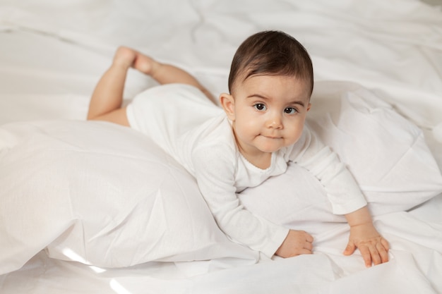 La bambina sveglia in un body bianco si trova a letto con biancheria di cotone. Foto di alta qualità.