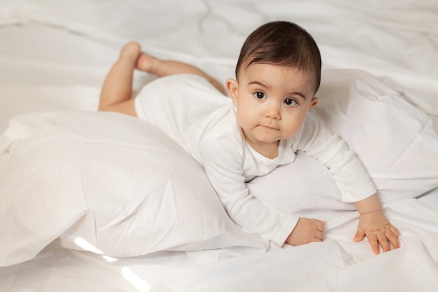 La bambina sveglia in un body bianco si trova a letto con biancheria di cotone. Foto di alta qualità.