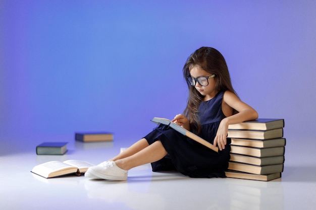 La bambina sveglia in vestito blu scuro e occhiali sta leggendo e studiando il libro. bambino e molti libri. Bambini e istruzione.