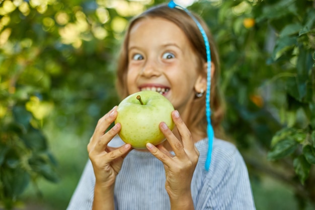 La bambina sveglia mangia la mela verde nel giardino domestico all'aperto, bambino felice