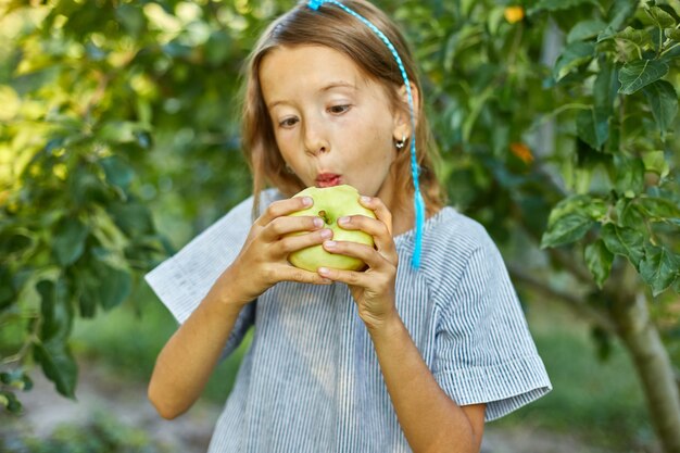 La bambina sveglia mangia la mela verde nel giardino domestico all'aperto, bambino felice