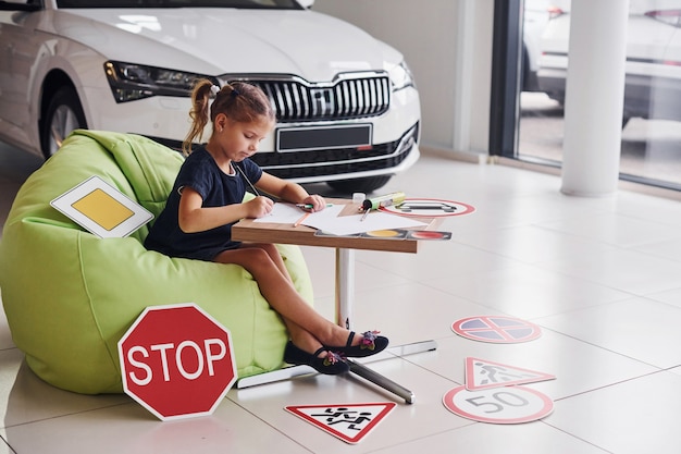 La bambina sveglia si siede sulla sedia verde morbida vicino al tavolo con fogli di carta e matita. Vicino alla moderna segnaletica stradale e automobilistica sul pavimento.