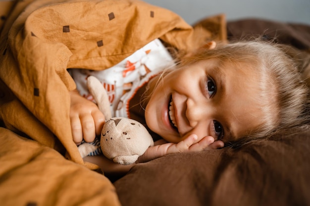La bambina sveglia si trova accogliente nel letto coperta con una coperta sopra la sua testa che sorride con lo spazio della copia
