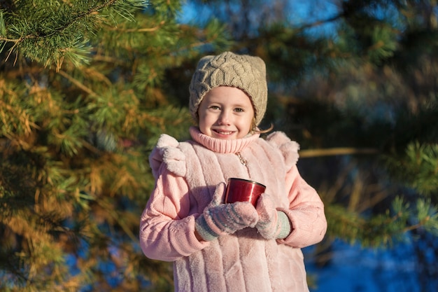 La bambina sveglia sta tenendo una tazza con tè caldo nella foresta di inverno. Infanzia felice. Bambini all'aperto Divertimento invernale concetto di vacanza