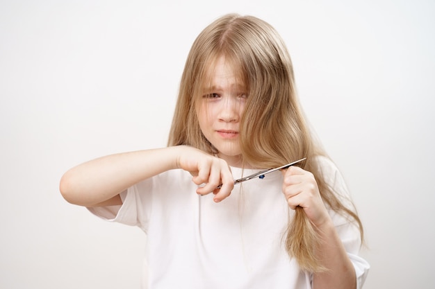 La bambina triste taglia i capelli lunghi con le forbici e ha paura su uno sfondo bianco. Taglio di capelli alla moda per il bambino. Parrucchiere. scherzi dei bambini. taglio di capelli