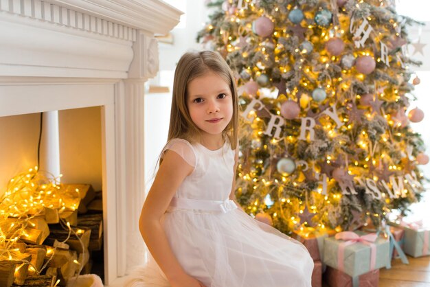 la bambina, vestita con un bellissimo abito bianco alla moda, posa vicino all'albero di Natale.