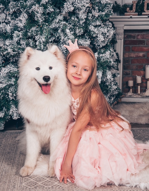 La bella bambina bionda sorride con un divertente cane Samoiedo bianco vicino all'albero di Capodanno.