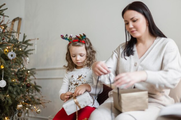 La bella bambina con la mamma si siede sul divano vicino all'albero di Natale e impacchetta i regali per Natale e Capodanno