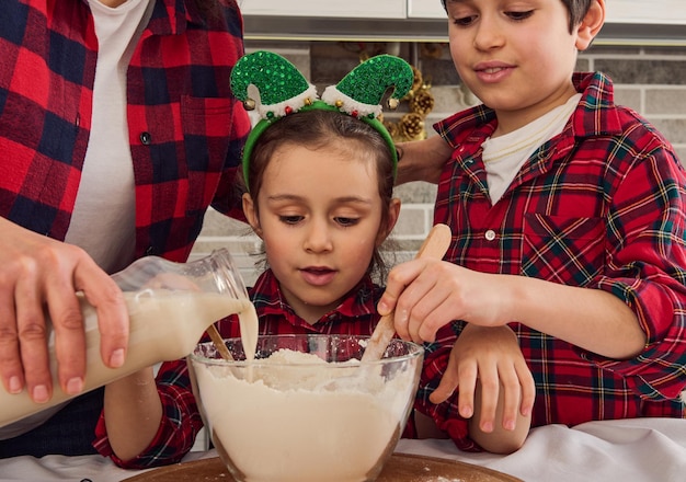 La bella bambina si siede tra la sua amorevole madre che versa del latte in una ciotola di farina e il suo allegro fratello maggiore che mescola e impasta gli ingredienti secchi mentre prepara l'impasto dei biscotti di Natale