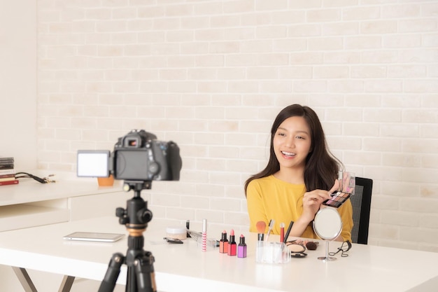 La bella blogger asiatica che indossa una maglietta gialla sta mostrando come truccarsi e usare i cosmetici Sta girando un video di se stesso usando una fotocamera DSLR su un treppiede