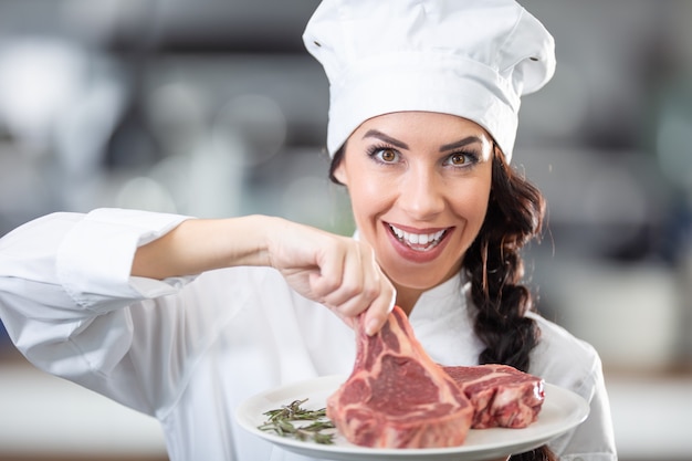 La bella chef donna in uniforme e cappello tiene un piatto con bistecche crude ed erbe fresche in cucina.