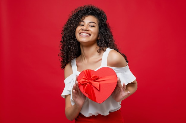 La bella donna africana felice che tiene il cuore ha modellato il contenitore di regalo del biglietto di S. Valentino isolato su rosso