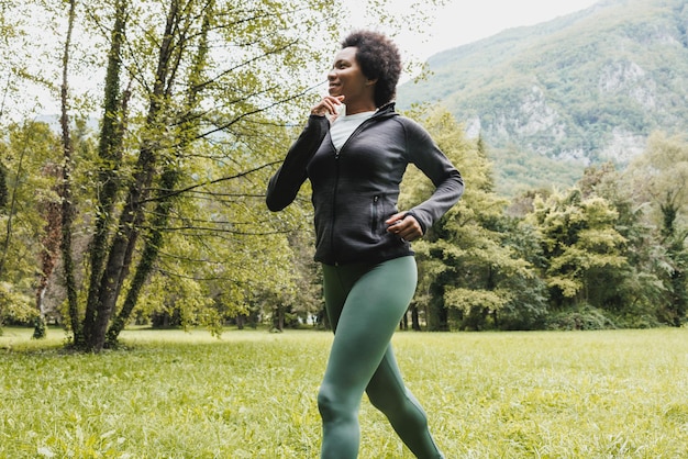 La bella donna afroamericana matura sorridente sta facendo jogging attraverso l'erba verde vicino al paesaggio di montagna.