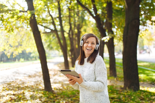 La bella donna allegra dai capelli castani felice in maglione bianco con un tablet che ascolta musica nelle cuffie bianche nel parco autunnale in una giornata calda. Autunno in città.