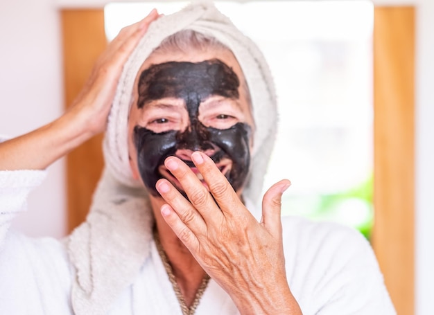 La bella donna anziana sfocata con una maschera di carbone facciale detox fatta in casa sorridente con la mano sulla bocca si prende cura del concetto di pelle