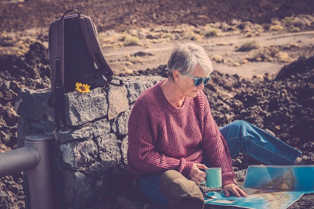 La bella donna anziana si siede all'aperto durante il viaggio di trekking con lo zaino e la mappa cartacea godendosi la natura e lo stile di vita alternativo con libertà e sentimento con il percorso del mondo