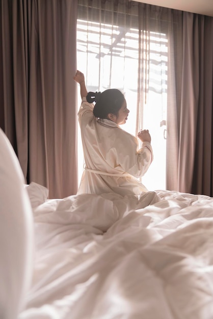 La bella donna asiatica del vestito bianco che allunga la mattina sveglia la freschezza e il momento del fine settimana spensierato nell'accogliente camera da letto interna con il concetto della casa di stile di vita del fondo della luce del sole e della tenda bianca