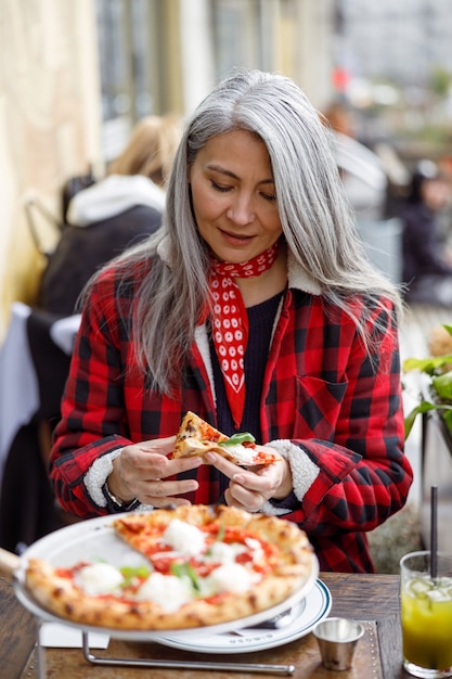 La bella donna asiatica matura sta mangiando la pizza nella terrazza del caffè