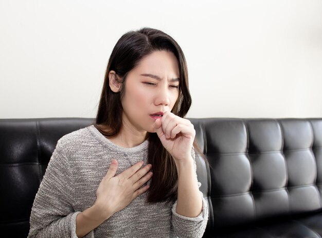 La bella donna asiatica seduta in soggiorno è tosse e usando la mano tocca il petto Concetto di assistenza sanitaria