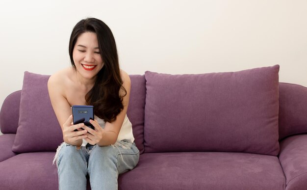 La bella donna asiatica seduta sul divano usa lo smartphone per chattare o fare acquisti online o sui social media durante la sua vacanza con felicità e sorriso