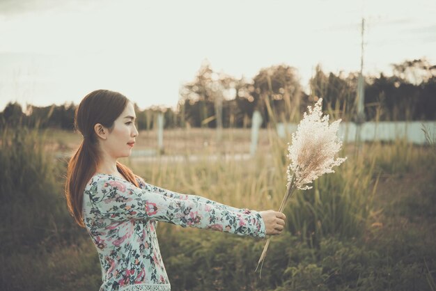 La bella donna asiatica tiene il fiore dell'erba nei campi con la luce solare intensa è molto felicestile vintagetono scuro al tramonto