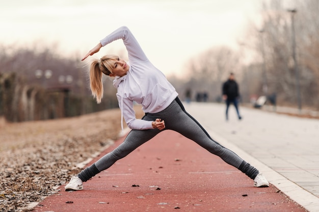 La bella donna bionda caucasica in abiti sportivi che fanno l'allungamento si esercita prima di correre all'aperto.