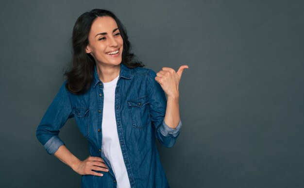 La bella donna castana alla moda moderna alla moda in vestiti del denim sta posando sui precedenti grigi