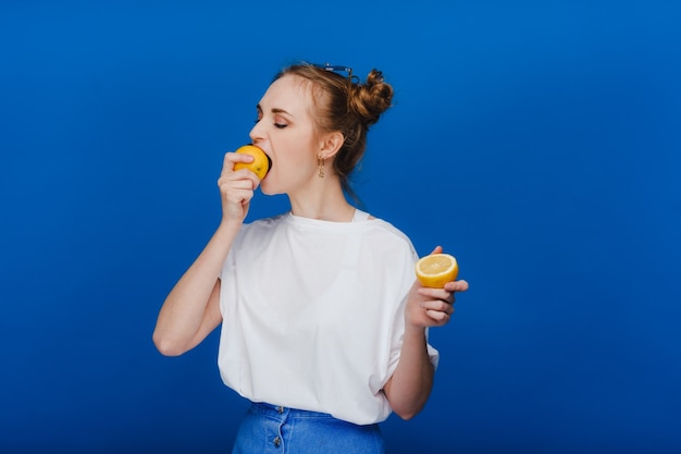La bella donna castana prende un morso di un limone. Ragazza su sfondo blu.