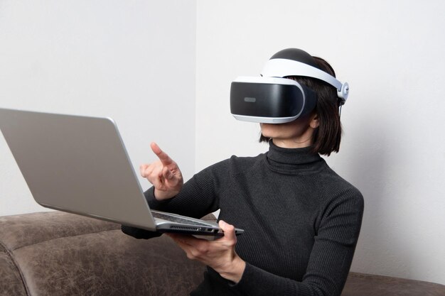 La bella donna che utilizza l'auricolare vr mostra qualcosa nel laptop, chatta con gli amici praticamente a casa. Il concetto di universo virtuale. Stile di vita nello spazio virtuale. Apprendimento virtuale online