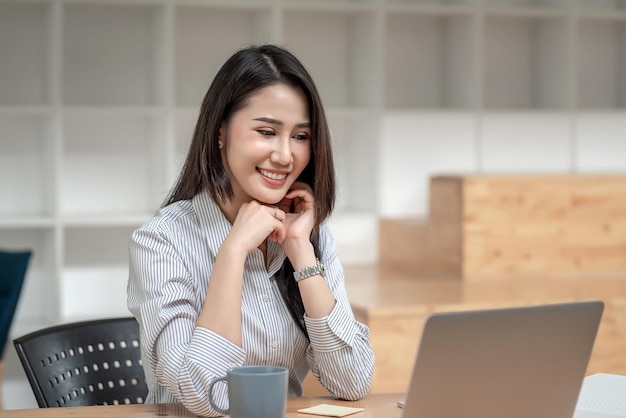 La bella donna d'affari asiatica sorridente è felice di sedersi e guardare il laptop in ufficio.