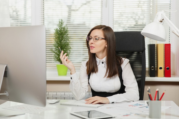 La bella donna d'affari dai capelli castani seria e assorta in giacca e occhiali seduta alla scrivania con tablet, lavorando al computer con monitor moderno con documenti in ufficio leggero, guardando da parte