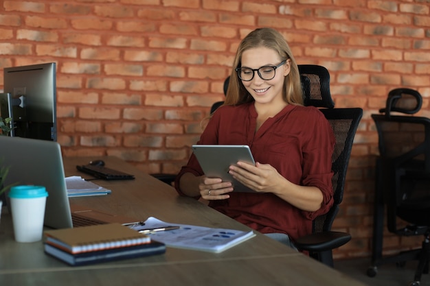 La bella donna d'affari sta lavorando utilizzando la tavoletta digitale mentre è seduta in ufficio creativo.