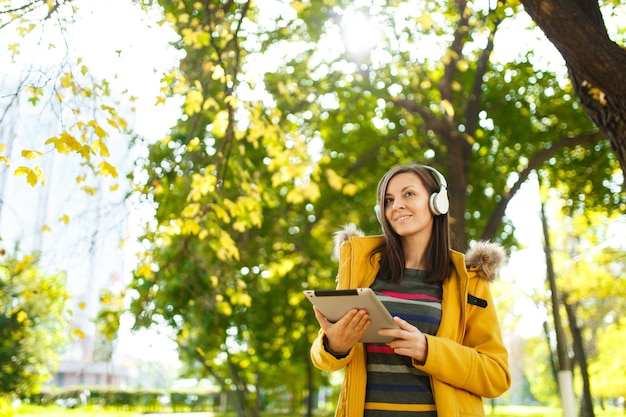 La bella donna dai capelli castani allegra felice in un cappotto giallo e maniche lunghe a righe esulta con un tablet in mano e cuffie bianche nel parco cittadino autunnale in una giornata calda. Foglie d'oro autunnali.