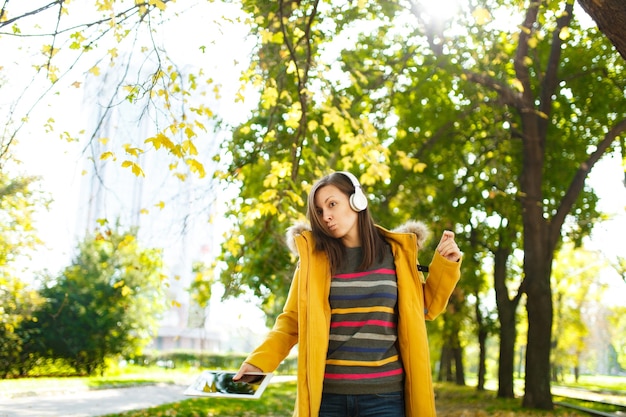 La bella donna dai capelli castani allegra felice in un cappotto giallo e maniche lunghe a righe esulta con un tablet in mano e cuffie bianche nel parco cittadino autunnale in una giornata calda. Foglie d'oro autunnali.