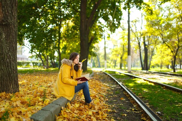 La bella donna dai capelli castani felice in un cappotto giallo e jeans si siede da sola nel parco vicino ai binari del tram e legge un libro nella calda giornata autunnale. Foglie gialle autunnali.