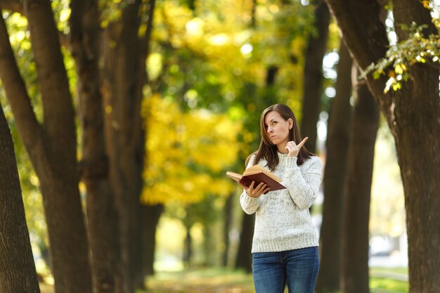 La bella donna dai capelli castani sorridente felice in maglione bianco in piedi con un libro rosso nel parco cittadino autunnale in una giornata calda. Foglie d'oro autunnali. Concetto di lettura.