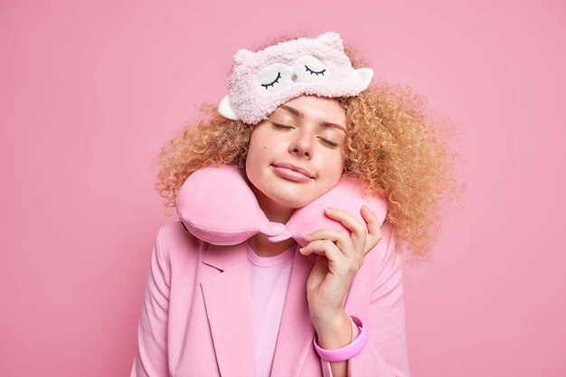 La bella donna dai capelli ricci vede sogni piacevoli mentre fa un pisolino durante il giorno tiene gli occhi chiusi indossa un cuscino per il collo bendato per un riposo confortevole gode di un'atmosfera calma isolata sul muro rosa