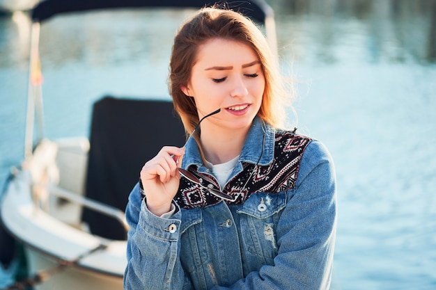 La bella donna del primo piano con capelli biondi tiene gli occhiali rotondi in sua mano in una giacca del denim, il giorno di estate sulla via nel porto