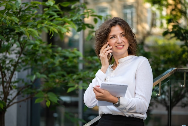 La bella donna di affari sta parlando su uno smartphone. Ha in mano un taccuino.