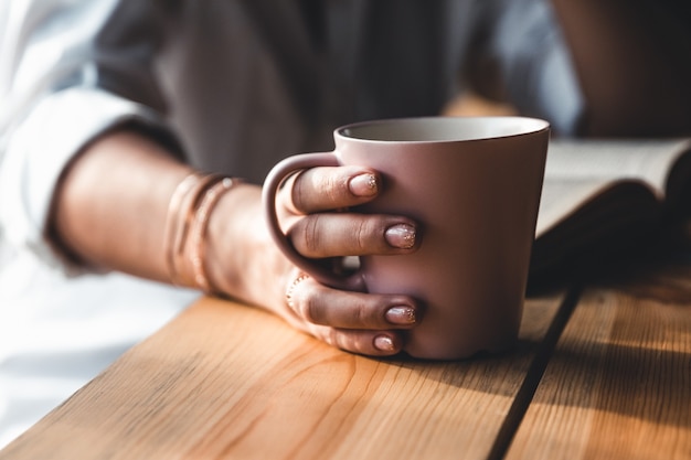 La bella donna di mattina beve il caffè e legge un vecchio libro in una camicia bianca. Educazione, bere.