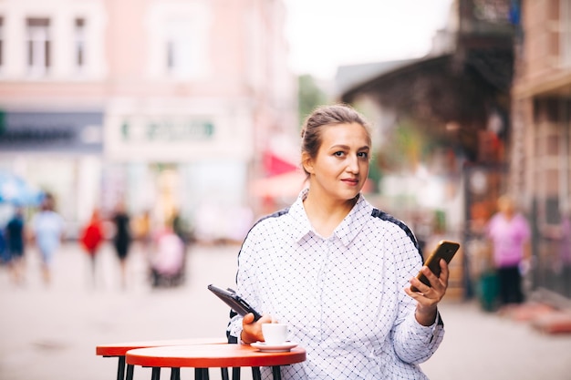 La bella donna è in piedi al tavolo in un caffè di strada con in mano tablet e smartphone