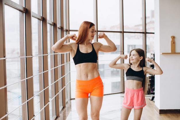 La bella donna forte e la piccola figlia affascinante stanno mostrando il loro bicipite e stanno sorridendo mentre si allenano in palestra